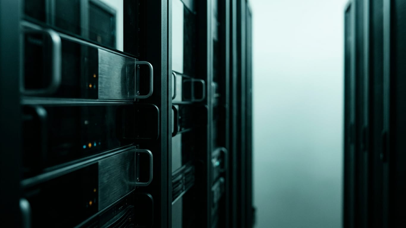 Illustration for this article: Hinton urges pre-release review for advanced AI. Rows of dark unbranded rack equipment receding down a narrow aisle, lit only by their own indicator glow.
